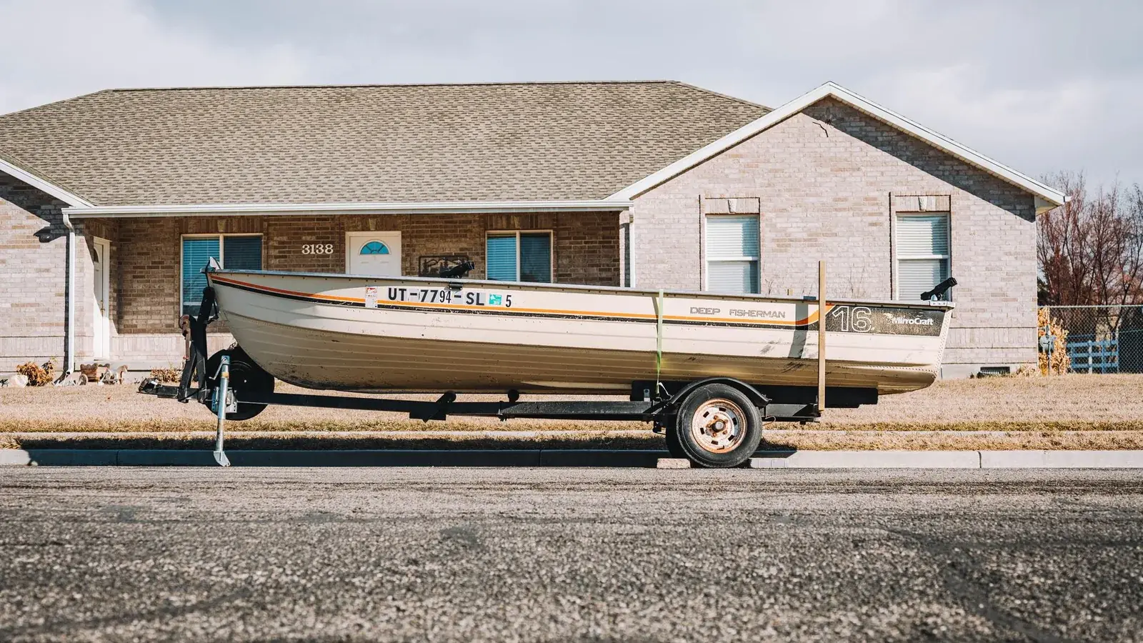 A sixteen foot aluminum fishing boat on a single-axle trailer parked at the curb outside a house, tongue jack down